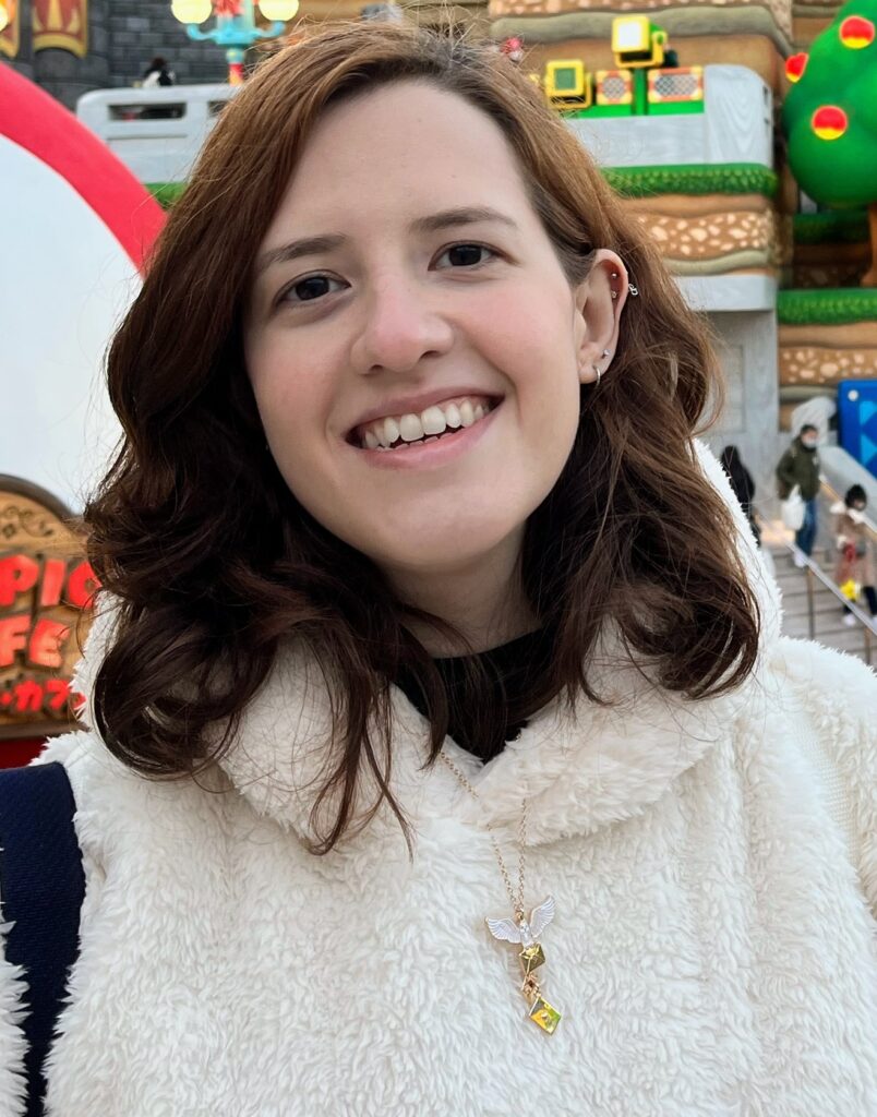 Imagem da professora Bruna sorrindo. Bruna tem pele branca, cabelos na altura do ombro ruivo. Está vestindo uma blusa de frio na cor bege. Ao fundo da imagem um fundo que se parece ao cenário de um jogo, com cores branca, vermelha, verde e marrom.