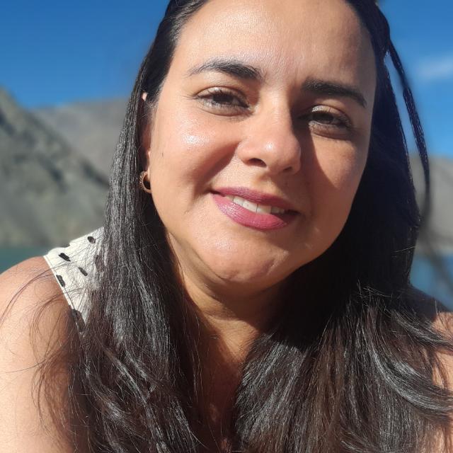 Imagem da professora Kamila sorrindo. Ela tem pele branca, cabelos lisos longos, e no fundo de sua imagem há um céu azul e montanhas.