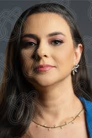 Kamila é uma mulher de pele clara, cabelos castanhos escuros e longos caindo sobre os ombros. Ela está olhando diretamente para a câmera, com uma maquiagem nos olhos em tons de rosa e marrom e batom rosa nos lábios. Veste uma roupa azul (apenas o colarinho e parte do ombro são visíveis), um colar fino com pequenas folhas pontiagudas e um brinco dourado em forma de flor na orelha direita visível. O fundo é cinza escuro, com um padrão celta entrelaçado, semitransparente, sobreposto à imagem, uma marca d'água do ICMC.