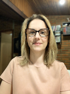 A professora Mariza tem pele branca, cabelos loiros na altura dos ombros e óculos de aro grosso marrom tartaruga, está olhando diretamente para a câmera. Ela veste uma blusa cor-de-rosa pêssego de manga curta e está em um ambiente interno. A iluminação é suave e o fundo desfocado revela uma parede de tijolos à vista e algumas prateleiras ou objetos no canto superior direito.